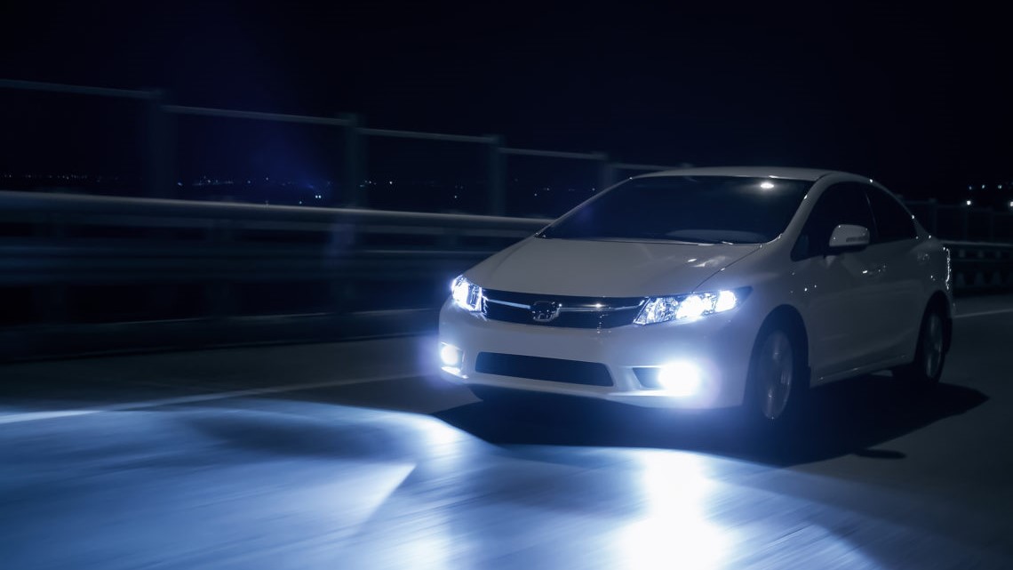 A white car driving on a bridge at night, its headlights illuminating the path ahead.