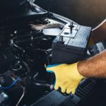 A person wearing yellow gloves installs a new car battery under the hood of a vehicle in a darkened garage.