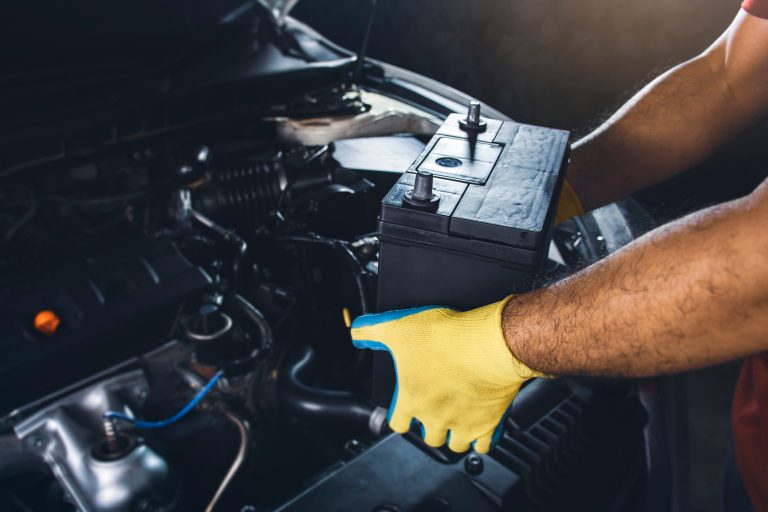 A person wearing yellow gloves installs a new car battery under the hood of a vehicle in a darkened garage.
