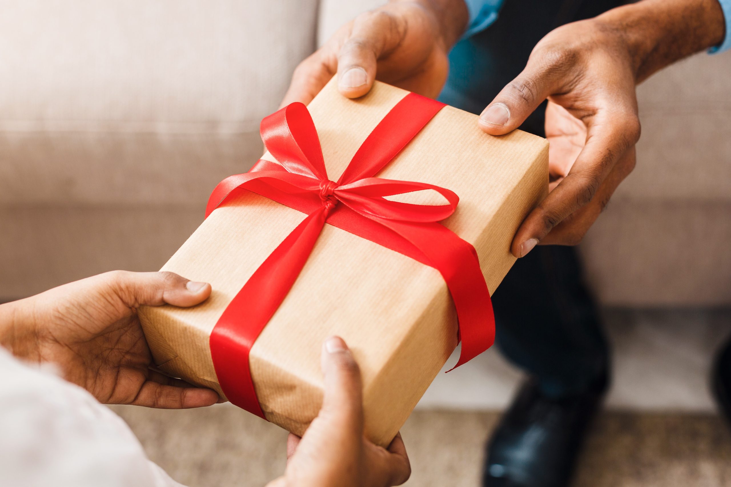 Two people exchanging a gift wrapped in brown paper with a red ribbon. only their hands are visible.