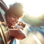 A person with curly hair smiles while leaning out of a car window on a sunlit road.