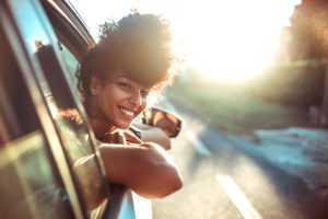 A person with curly hair smiles while leaning out of a car window on a sunlit road.