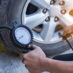 A person checking the air pressure of a car tire using a pressure gauge tool in daylight.