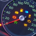 Close-up of a car dashboard displaying illuminated warning lights, including ABS, fuel, high beam, handbrake, engine, and others—essential for understanding car warnings—with the speedometer reading at 0 km/h.