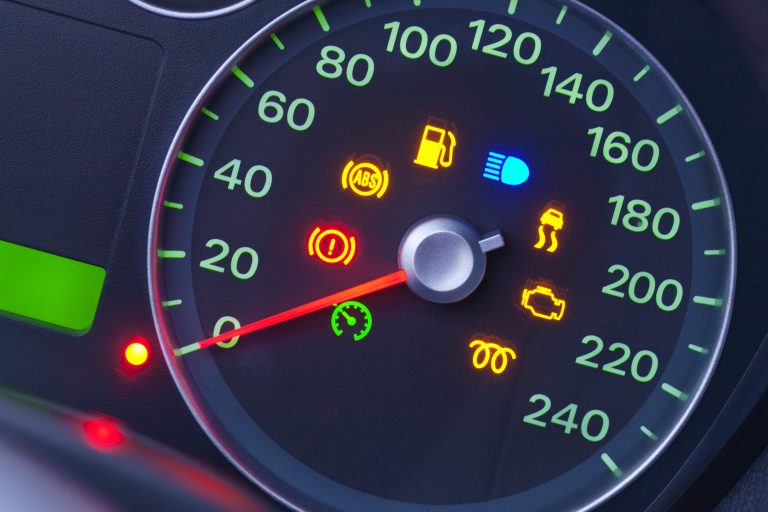 Close-up of a car dashboard displaying illuminated warning lights, including ABS, fuel, high beam, handbrake, engine, and others—essential for understanding car warnings—with the speedometer reading at 0 km/h.