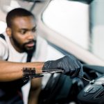 Young handsome concentrated African American man using cleaning brush and removing dust from car air conditioning vent grill.