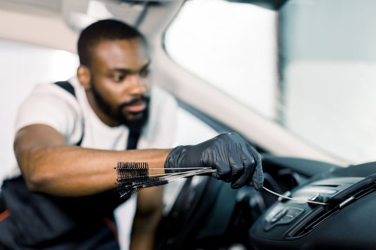 Young handsome concentrated African American man using cleaning brush and removing dust from car air conditioning vent grill.